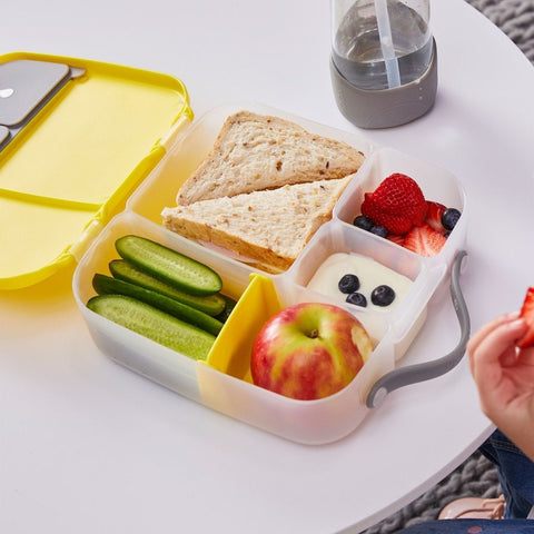 The B.Box Lunch Box by B.Box, styled like a bento box with leak-proof silicone seals, features compartments for sandwich halves, cucumber slices, yogurt with blueberries, strawberries, and a red apple. On the white table beside it is a reusable water bottle and someone enjoying a strawberry.