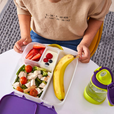 A person sits at a table with an open B.Box Lunch Box from B.Box in front of them. The flexible lunch box has compartments filled with strawberries, a yogurt parfait with berries, chopped salad with chicken, and a banana. A green and purple water bottle is next to the lunch container.