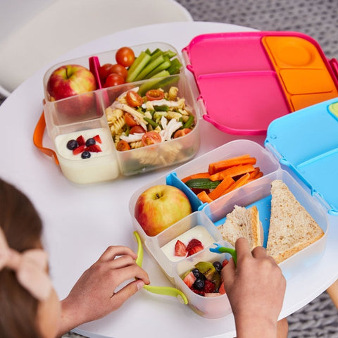 On a white table, two B.Box Lunch Boxes from B.Box are filled with a variety of healthy items. The lunchbox in the foreground contains a sandwich, vegetable sticks, an apple, yogurt with berries, and fruit salad. Equipped with leak-proof silicone seals, a child is holding a fork, ready to eat.