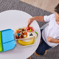 A child sits at a small white table, eating from a B.Box Lunch Box by B.Box. The lunchbox contains slices of watermelon, blueberries, and other small food items, along with a banana on the side. The child is reaching for a piece of food with their hand, enjoying the freshness from the whole foods in the B.Box Lunch Box.