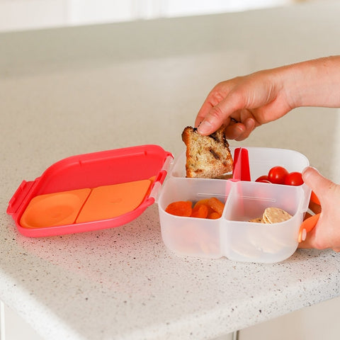 A person holding a small toasted sandwich piece over the B.Box Mini Lunch Box by B.Box, which features sections for cherry tomatoes, carrot slices, and a small dessert, on a speckled white countertop. The container has leak-proof compartments and a red lid.