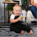 A happy baby wearing a white shirt and black overalls sits on a striped rug, holding a Baby Einstein Grip & Spin Oball Rattle Toy from Baby Einstein. The baby is smiling and looking forward. In the background, there is a person partially visible sitting on a chair and a small table with plants and books.