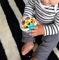 A baby wearing a striped shirt and gray pants sits on a black-and-white striped rug, grasping a Baby Einstein Grip & Spin Oball Rattle Toy with its vibrant colored sections inside. The baby's barefoot is visible, and their hands are engrossed with the toy.