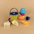 A collection of vibrant beach toys is arranged on a beige surface. It features a green bucket with a blue handle, a yellow rake, a white scoop, a blue and purple whale mold, an orange fish mold, a purple shell mold, and a yellow toy boat—all ideal for filling your Battat Beach Wagon with Toys by Battat or crafting exquisite sandcastles.