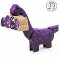 A wooden dinosaur toy, mainly purple with lighter purple spots, holds a segmented wooden caterpillar in its mouth. The caterpillar's segments are connected in a curved line. This piece is part of the Brio World Dinosaur Adventure Set by Brio, which includes various dinosaur-themed playset elements.