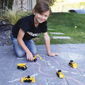 A young boy wearing a black "Caterpillar" t-shirt is playing outdoors on a paved surface with his CAT Little Machines 5 Pack, which includes several small, yellow construction vehicles like a dump truck, excavator, and bulldozer. He is smiling and looking at the camera while role-playing. A garden is visible in the background.