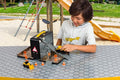 A young boy wearing a light-colored hoodie plays with the CAT Little Machines Store-n-Go Playset at an outdoor playground. The set includes miniature construction vehicles, road signs, and cones arranged on a patterned table. In the background, a slide and climbing structure are visible.