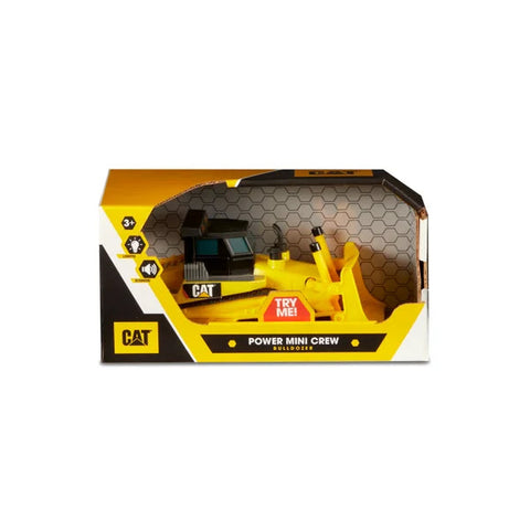 A yellow toy bulldozer, branded as the CAT Power Mini Crew - Bulldozer, is packaged in a black and yellow box. The packaging features a "Try Me!" button, realistic lights and sounds, and icons indicating it's suitable for ages 3 and up. Part of the push-powered vehicle is visible through a clear plastic window.