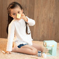 A young girl with braided hair sits on a carpeted floor against a wooden wall, holding a toy cup to her mouth. Beside her is the Classic World Coffee Maker by Classic World, along with a small milk carton and mug, suggesting she's pretending to make coffee. She wears a white long-sleeved shirt and denim shorts.