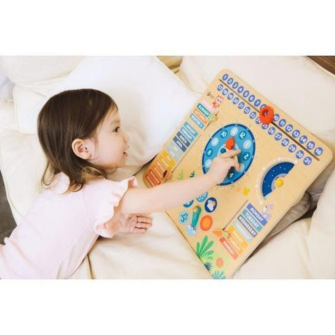 A young child with brown hair is lying on a white cushion and engaging with the Classic World Forest Calendar, an educational wooden activity board from Classic World. This learning tool, crafted from natural wood, features colorful dials, gears, and letters designed to enhance learning through play.