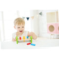 A young child with blonde hair is joyfully playing with the Classic World Rocking Pounding Bench, pushing colored pegs through holes in the natural wood. The toy is placed on a white table. In the background, there's a white room adorned with triangular bunting and a colorful storage unit, fostering excellent hand-eye coordination.