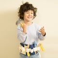 A young child with curly hair and a light blue shirt smiles while holding a toy hammer. They proudly display their Classic World Tool Belt, showcasing carpentry tools for kids such as a wrench, screwdriver, and tape measure. The neutral light background enhances this moment of creative play.