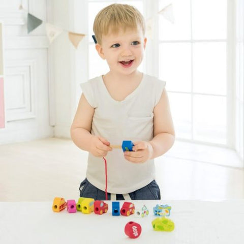 A young child smiles while playing with the Classic World Traffic Beads from Classic World on a white table. The child is threading a red string through one of the colorful, block-shaped toys and wooden vehicles. The background features a bright room with large windows and decorative bunting on the wall, fostering imaginative play.