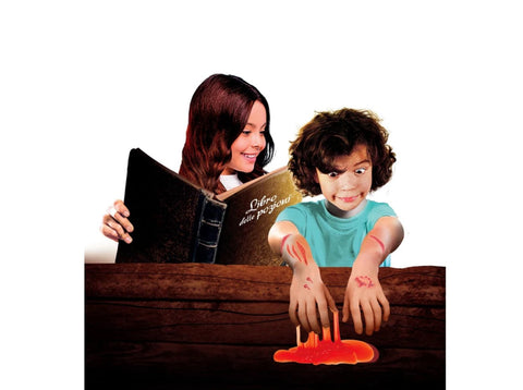 A child with a mischievous smile is squeezing a red substance between their fingers, creating a gooey mess on a wooden table. Behind them, an adult with long hair is holding an open book titled "Libro de poción" and looking at the child as if curating spooky slime experiments from the Clementoni Science & Play - Fun Vampire and Blood Kit by Clementoni.