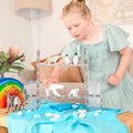 A young girl in a light blue dress plays with plastic animal figurines, including a cheetah, penguins, polar bears, and reindeer. She arranges them on Connetix Clear Rectangles from the 12-piece pack atop a blue cloth, with magnetic tiles and a colorful rainbow toy nearby. White pebbles accentuate her creative STEAM learning setup.