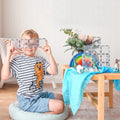 A young boy wearing a striped shirt with an orange robot design sits cross-legged on a cushion. He holds Connetix Clear Rectangle Pack (12pc) over his eyes, smiling. To his right is a wooden table filled with toys perfect for STEAM learning, including magnetic tiles, a rainbow stack, animals, and fabric.