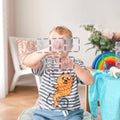 A child dressed in a striped shirt featuring an orange, bicycle-riding cartoon character proudly holds up a structure made with the Connetix Clear Rectangle Pack 12pc from Connetix. Seated in a bright room adorned with a decorative chair, plants, and colorful toys, they are fully immersed in STEAM learning through play.