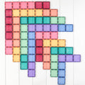 A collection of colorful Connetix shapes arranged in overlapping rectangular patterns on a white wooden surface. The tiles, from the Connetix Pastel Rectangle Pack 24pc, feature strong magnets and come in various pastel colors including red, pink, orange, yellow, green, blue, and purple. They create visually appealing geometric designs that are perfect for STEAM learning.