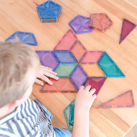 A child, wearing a striped shirt, is focused on arranging colorful magnetic tiles from the Connetix Pastel Shape Expansion Pack 48pc on a wooden table. The child creatively organizes the Connetix tiles, which come in various shapes such as triangles, squares, and hexagons, into a star-like pattern.