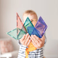 A child in a striped shirt proudly holds up an array of colorful translucent magnetic tile shapes from the Connetix Pastel Shape Expansion Pack 48pc, including triangles, squares, and semicircles. The background is blurred, featuring a light-colored wall and a piece of furniture.