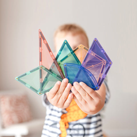 A child in a striped shirt proudly holds up an array of colorful translucent magnetic tile shapes from the Connetix Pastel Shape Expansion Pack 48pc, including triangles, squares, and semicircles. The background is blurred, featuring a light-colored wall and a piece of furniture.