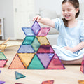 A young girl in a blue dress smiles while playing with the Connetix Pastel Shape Expansion Pack 48pc, arranging the colorful geometric magnetic tiles into a pyramid structure topped with a star. She is seated on a wooden floor in a bright room, delighting in her growing mathematical understandings.