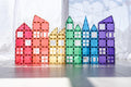 A series of colorful Connetix creations made from the Connetix Rainbow Mega Pack 212 pc magnet tiles arranged to resemble a row of houses in the shape of rectangles and triangles. The tiles are red, orange, yellow, green, blue, and purple, creating a rainbow effect against a bright, blurred background.