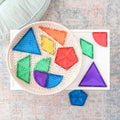 A variety of colorful geometric shapes, including triangles, squares, and hexagons from the Connetix Rainbow Shape Expansion Pack 36pc by Connetix, are placed in a woven basket and on a white table. The shapes come in red, orange, yellow, green, blue, and purple for creative play and STEAM learning. A blue shape is on the table.