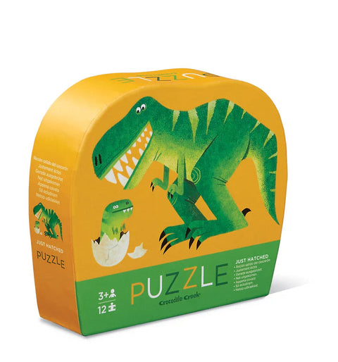 A vibrant, curved packaging box that houses the Crocodile Creek 12pc Mini Puzzle - Just Hatched. The box is mostly yellow and orange and showcases a fun green T-Rex dinosaur alongside a hatchling emerging from an egg. This jigsaw puzzle by Crocodile Creek fosters hand-eye coordination and is designed for children ages 3 and up.