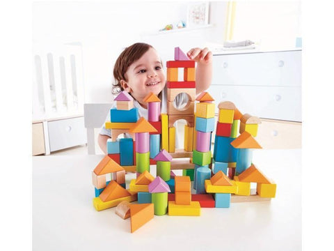 A joyful child with brown hair is engrossed in playing with the HAPE 101pcs Blocks set, expertly building a large structure on the table using sturdy beech blocks. The bright room is beautifully decorated with white furniture, including a crib and a dresser in the background. This engaging activity aids in developing dexterity.