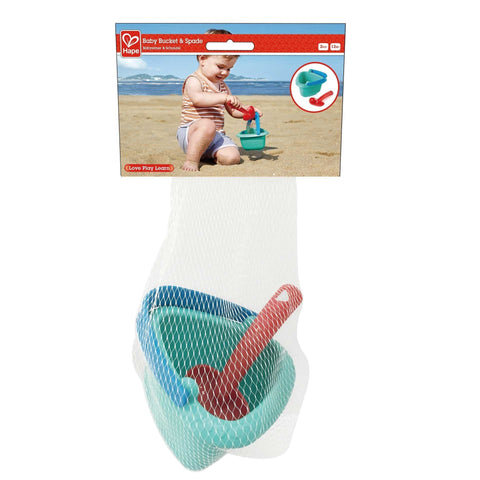 The HAPE Baby Bucket & Spade, perfect as a beginner's playset, is showcased in its packaging. The set includes a blue bucket and a red spade, with packaging featuring an image of a child playing with the beach set on the shore. Both items are neatly contained in a white mesh bag.