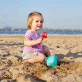 A young child with light hair, wearing a purple shirt and grey shorts, sits on a sandy beach holding the red scooper from their first HAPE Baby Bucket & Spade playset. A blue bucket from the HAPE brand is placed in front of them. The child is smiling with the ocean visible in the background under a clear blue sky.