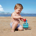 A toddler with light hair sits on a sandy beach, playing with their first playset: the HAPE Baby Bucket & Spade, which includes a red and blue shovel and a small green bucket. The child is wearing a striped tank top and gray shorts, while the background features the ocean and a distant shoreline under a clear blue sky.