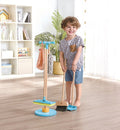 A young child with light brown hair, wearing a gray shirt with a brown pocket and blue shorts, smiles while holding the HAPE Broom & Swiffer Cleaning Stand from HAPE. The child stands in a room with wooden flooring, a floor-to-ceiling window, and a shelf adorned with various items and decorations.