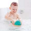 A smiling baby enjoys bath time, playing with the HAPE Bubble Maker Whale in a bathtub filled with bubbles. The baby holds the blue, orange, and white striped toy from HAPE, making water splash around as they giggle joyfully. Bubbles float through the air, adding to the fun in the clean white bathtub.