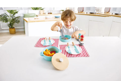 A child with light brown hair sits at a white table set with two turquoise bowls, peach slices in one, red and white checkered placemats, wooden utensils, and a charming HAPE Cook & Serve Set from the brand HAPE. The child is scooping food from a bowl. The background features a modern kitchen with plants and appliances.