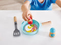 A child wearing a blue shirt is playing with the HAPE Cook & Serve Set by HAPE. The set includes a small frying pan with a toy fried egg and bacon, a grey spatula with a wooden handle, and a blue salt shaker. The child is holding a red item above the pan.