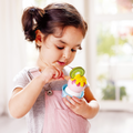 A young child in a light pink overall and gray shirt holds a toy from the HAPE Creamy Dreamy Ice Cream Corner set, topped with a kiwi slice. The child looks intently at the toy, using a small utensil to engage in imaginative pretend play. The background is bright and slightly blurred.