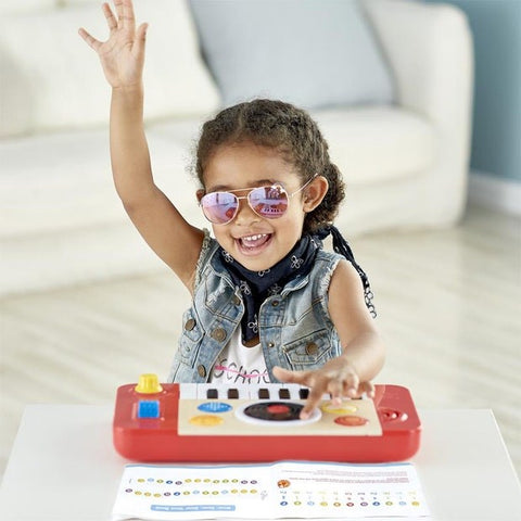 A young child with curly hair, wearing sunglasses and a denim vest, is grinning and raising one hand in the air while playing an epic keyboard solo on the colorful HAPE DJ Mix & Spin Studio toy keyboard placed on a white table. A booklet featuring various symbols lies open in front of the child.