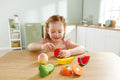 A young child with auburn hair is sitting at a wooden table in a bright kitchen, playing with the HAPE Farm Shop Fruit Playset. The colorful toy fruits from HAPE include an apple, banana, watermelon slice, kiwi, avocado, and orange slice. The child is holding a safe play knife from the set, pretending to cut the apple and developing fine motor skills.