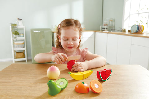 A young child with auburn hair is sitting at a wooden table in a bright kitchen, playing with the HAPE Farm Shop Fruit Playset. The colorful toy fruits from HAPE include an apple, banana, watermelon slice, kiwi, avocado, and orange slice. The child is holding a safe play knife from the set, pretending to cut the apple and developing fine motor skills.