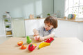 A young child is sitting at a table in a brightly lit kitchen, playing with the HAPE Farm Shop Fruit Playset by HAPE. The set includes a pear, tomato, apple, watermelon slices, and banana pieces. The child is honing their fine motor skills by holding and cutting a toy apple with the play knife included in the set. Shelves and dishes are visible in the background.