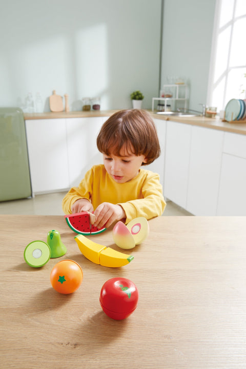 A young child sits at a wooden table in a brightly lit kitchen, playing with the HAPE Farm Shop Fruit Playset. The set includes colorful pieces like a watermelon slice, avocado, banana, pear, orange, and apple. Using the safe play knife to "cut" the fruit enhances fine motor skills. Dressed in a yellow long-sleeve shirt, the child is deeply focused on their HAPE toys.