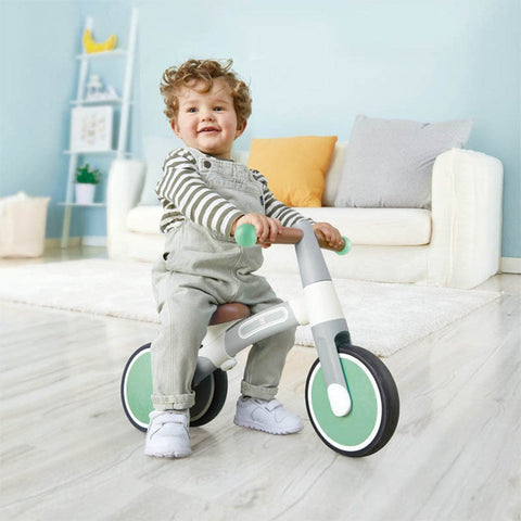 A toddler with curly hair is sitting on the HAPE My First Balance Bike - Green, a three-wheeled adjustable balance bike made by HAPE, indoors. The child is wearing a striped shirt and grey overalls, smiling with joy. The lightweight aluminium alloy design adds to the fun. The room has a white sofa, a small yellow pillow, and a shelf with decor in the background.