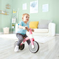 A toddler wearing a blue shirt and grey pants rides the HAPE My First Balance Bike Pink, featuring an adjustable design with a lightweight aluminium alloy frame, indoors. Behind the child is a white sofa adorned with grey and yellow cushions, next to a small white shelf, against pastel green walls decorated with artwork.