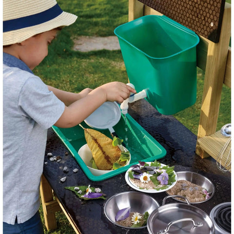 A young child wearing a sun hat enjoys outdoor play at the HAPE Outdoor Kitchen by HAPE. They are pouring water from a jug into a green basin filled with leaves and dirt. Several metal bowls with flowers, sand, and dirt are placed on the wooden toy playset's muddy surface.