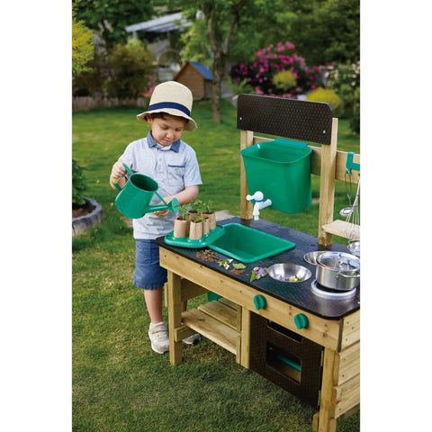 A young child wearing a straw hat and a light button-up shirt waters plants in small pots using a green watering can. The child is standing in a green garden beside the HAPE Outdoor Kitchen, featuring toy utensils and cooking accessories by HAPE, creating the perfect scene for outdoor play.