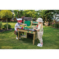 Two children are enjoying the outdoors with the HAPE Outdoor Kitchen by HAPE. One child, wearing a red and white hat and apron, is stirring a bowl on the counter of the wooden playset. The other child, wearing a light-colored hat, is observing and holding a spoon. Lush greenery provides the perfect backdrop for their outdoor play adventure.