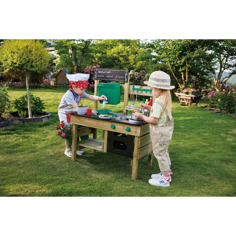 Two children are enjoying the outdoors with the HAPE Outdoor Kitchen by HAPE. One child, wearing a red and white hat and apron, is stirring a bowl on the counter of the wooden playset. The other child, wearing a light-colored hat, is observing and holding a spoon. Lush greenery provides the perfect backdrop for their outdoor play adventure.