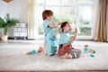 Two young children engage in role-play as budding hairstylists on a light-colored carpet in a sunlit room. One child, wearing a blue apron, pretends to style the other's hair, who smiles and sits with a cape around their neck. HAPE Super Stylish Hair Salon Set tools and accessories are scattered around, creating their own little hair salon.